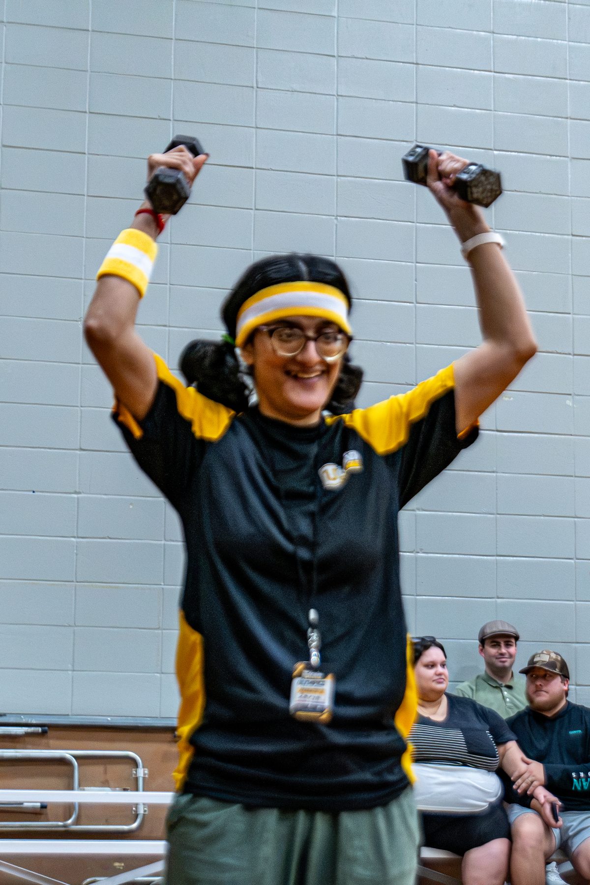 An athlete in a headband lifts two dumbbells overhead and smiles