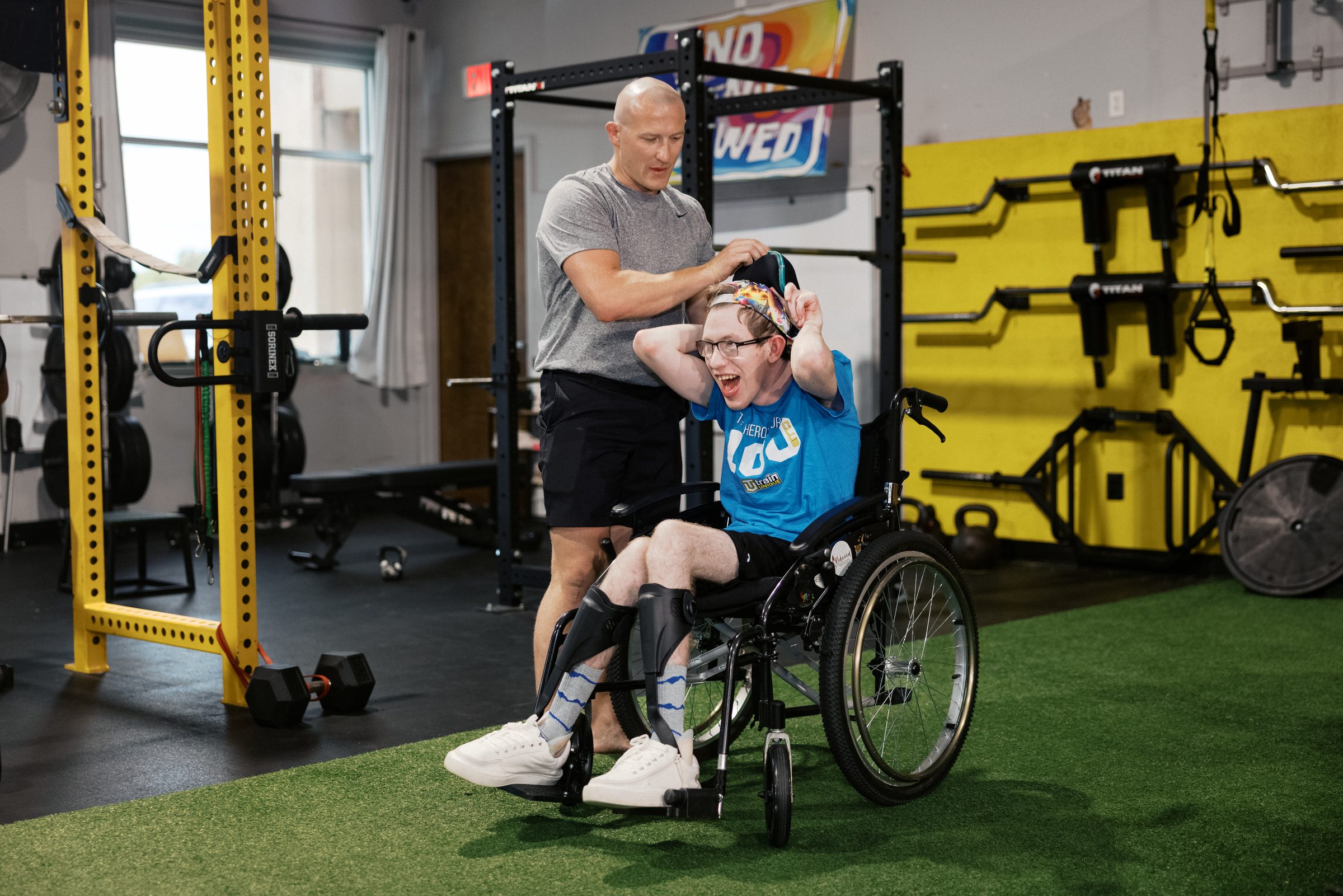 A coach stands behind an athlete in a wheelchair and adjusts his cap while the athlete, hands behind his head, laughs