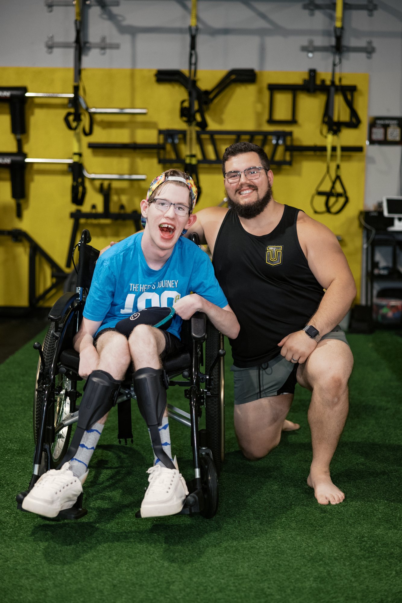 An athlete in a Hero's Journey 100 shirt laughs from his wheelchair while a coach kneels beside him, smiling